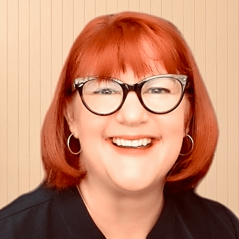 Amy Petre Hill headshot: A woman with red hair, glasses hooped earrings smiles and faces the camera