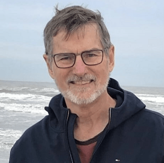 Bill Gaventa Headshot: Grey-haired man with glasses smiles in front of a beach.