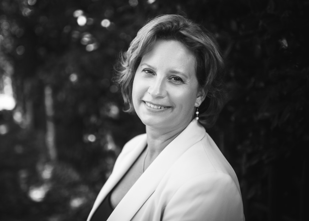 Black-and white photo of a smiling white woman (Karen) looking at the camera with medium-length hair, a pearl earing and a light-blazer.